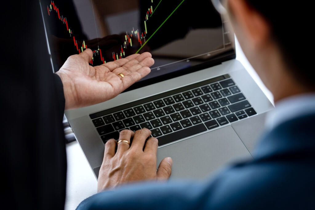 Imagem de uma equipe de negócios discutindo investimentos e estratégias de mercado em um ambiente de escritório. Os membros da equipe, em trajes profissionais, estão focados na análise de dados e gráficos do mercado de ações, que aparecem em uma tela à sua frente. A cena destaca a colaboração e a análise de dados financeiros para a tomada de decisões estratégicas em investimentos.